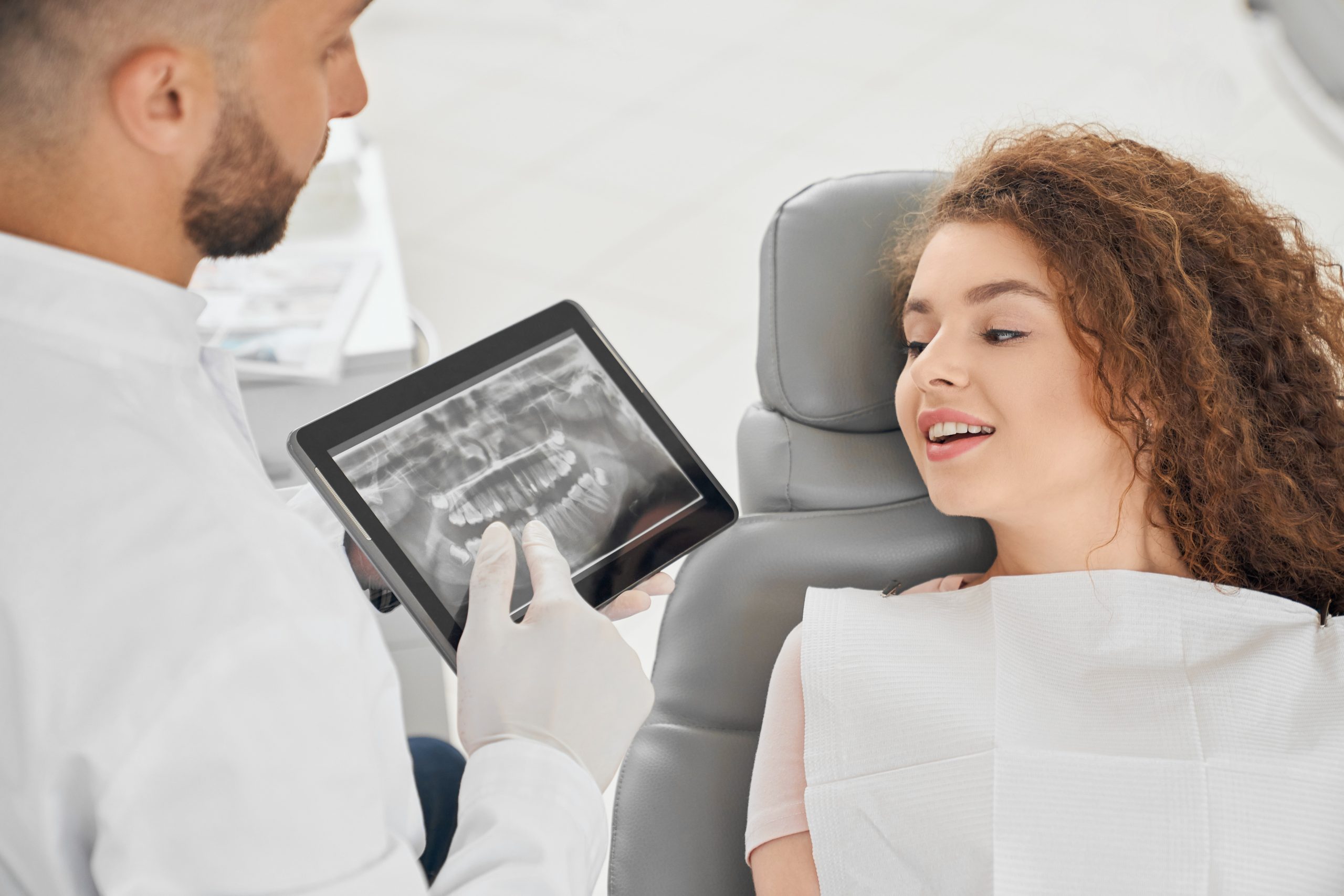 Front view of skilled male dentist in white uniform showing x ray picture of teeth to pretty woman. Female client sitting on chair and looking at tablet. Concept of treatment and health.